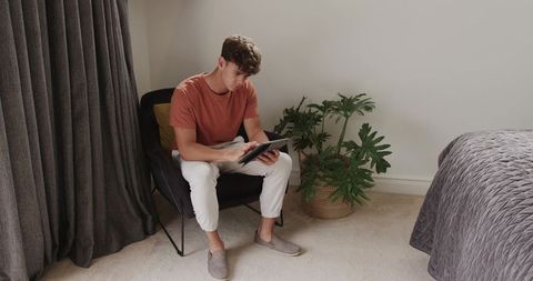 Young man using tablet with stylus in minimalist bedroom chair surrounded by plant