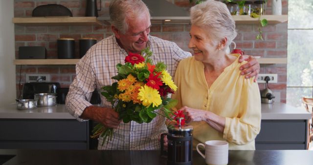 Happy senior caucasian husband giving flowers to his smiling wife in kitchen. Romance, love, senior lifestyle and togetherness