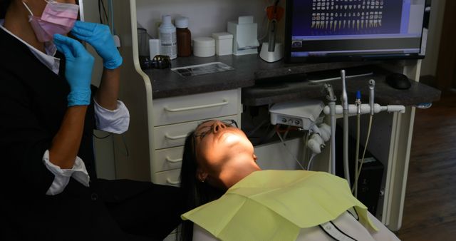 Woman lying on dental chair receiving dental examination. Dentist wearing gloves and mask preparing instruments. Bright overhead light illuminating patient's face. Computer screen showing dental images. Ideal for medical, dental health, and healthcare promotion materials.