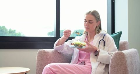 Female nurse enjoying healthy lunch break in lounge wearing pink scrubs and stethoscope