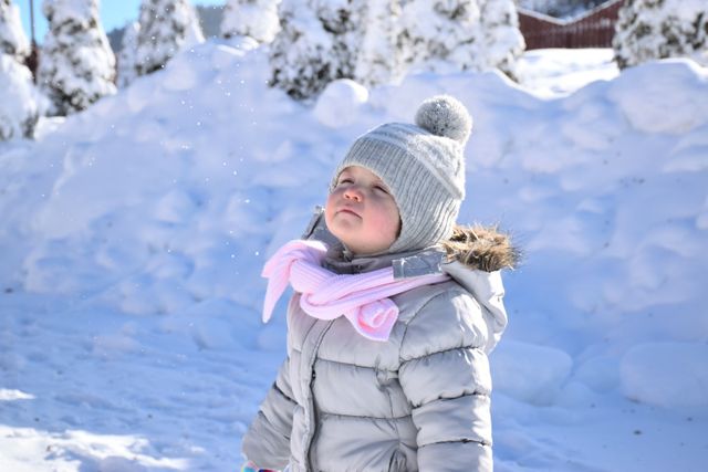 A young toddler dressed in a grey hat with a pom-pom and a pink scarf enjoying the snowy outdoors. This can be used for content related to winter activities, children's winter fashion, family adventures, or seasonal holiday themes.
