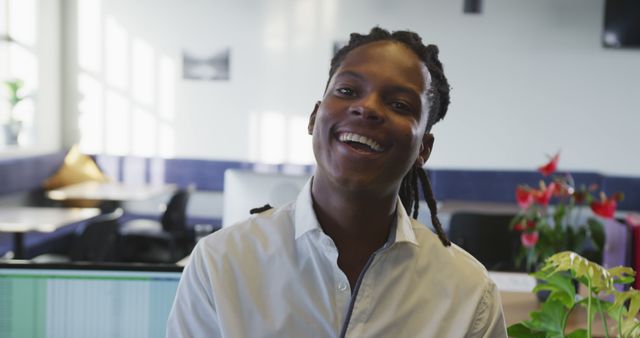 Man with dreadlocks smiling in a modern, well-lit office environment. Various workspaces and greenery in the background add to the professional yet relaxed atmosphere. Suitable for use in articles about workplace diversity, employee engagement, modern offices, and business professional environments.