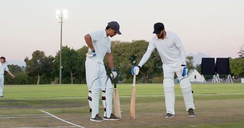 Two cricketers preparing at crease during dusk match with floodlight and protective gear