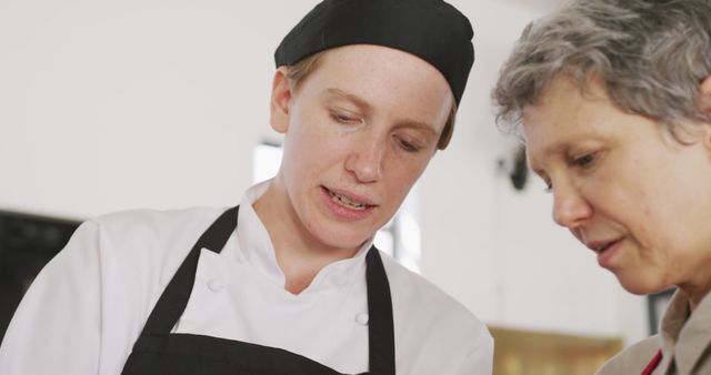 Professional chefs are seen discussing a recipe in a kitchen. The chef in the foreground listens attentively while another chef explains something. This image can be used for articles or advertisements related to culinary courses, restaurant teamwork, professional kitchen environments, or recipe development.