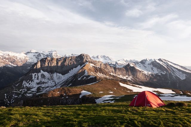 Showcasing a vibrant red tent set on a green mountain meadow with distant snowy peaks at twilight. Perfect for projects related to outdoor adventures, wilderness exploration, nature photography, camping gear promotions, and travel blogs.