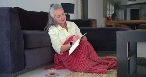 Mature woman writing in notebook on floor wrapped in red blanket in cozy living room