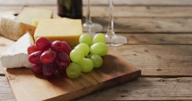 A well-organized cheese platter featuring various kinds of cheese and fresh red and green grapes is displayed on an elegant wooden board. There are also blurry wine glasses and a bottle of wine in the background, all set on a rustic wooden table. This could be used in food blogs, culinary magazines, wine and cheese tasting events, and party planning ideas.