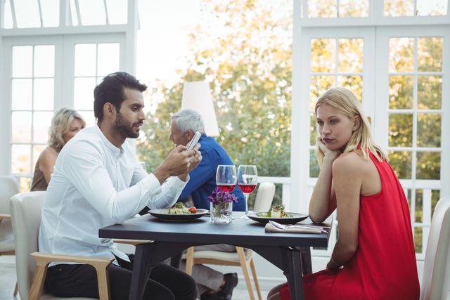 Couple sitting at a restaurant table, man focused on his smartphone while woman looks bored and uninterested. Ideal for illustrating themes of modern relationships, technology's impact on communication, and social disconnect. Suitable for articles, blogs, and advertisements related to dating, relationship advice, and technology use.