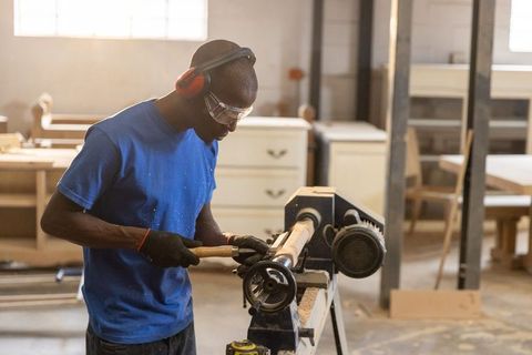 Woodturner carving wooden spindle on lathe in workshop wearing safety goggles, earmuffs