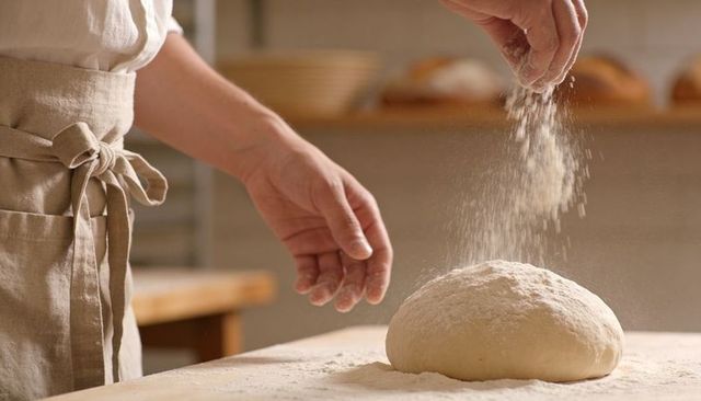 Baker sprinkling flour over artisan bread dough on floured wooden table, linen apron