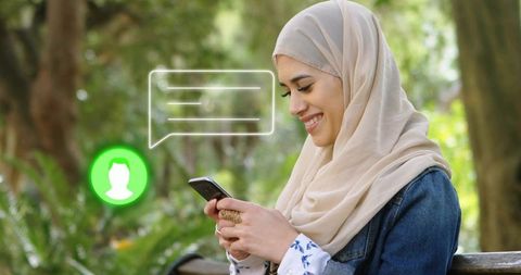 Young muslim woman wearing beige hijab messaging on smartphone while sitting on park bench