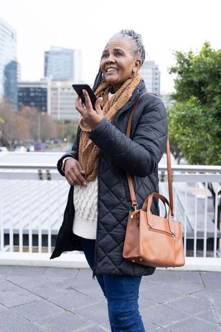 Senior woman holding smartphone on urban bridge wearing quilted coat and scarf