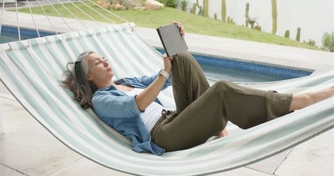 Senior woman reclining on striped hammock at poolside holding tablet and wristwatch