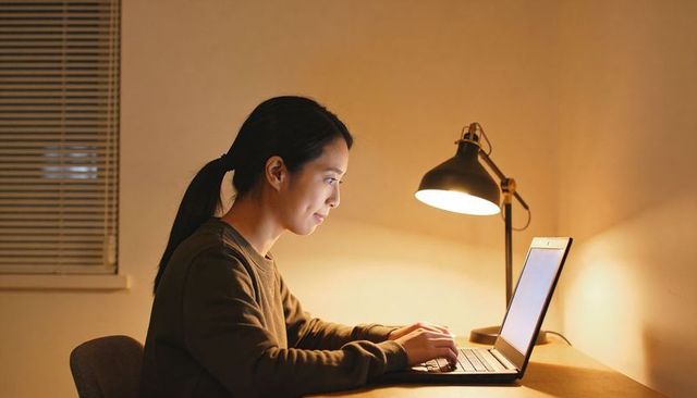 Asian woman working late on laptop at minimalist home office desk with warm lamp glow
