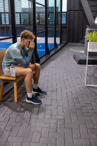 Exhausted young athlete sitting on bench beside padel court fencing covering face with hands
