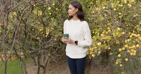 Young woman enjoying hot drink in lemon orchard wearing cream sweater and smartwatch