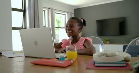 African american girl studying on laptop at home smiling while using headphones and books