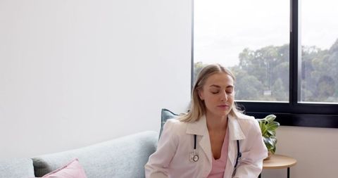 Female doctor resting on clinic sofa wearing white coat and stethoscope, calm wellness