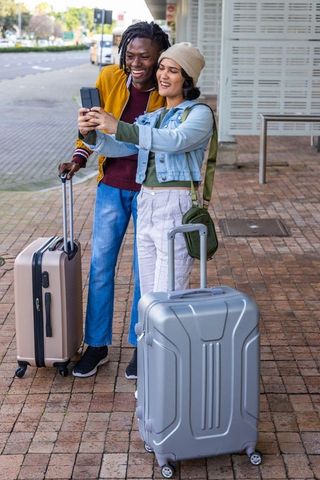 Multicultural couple taking selfie at urban transit stop with rolling luggage