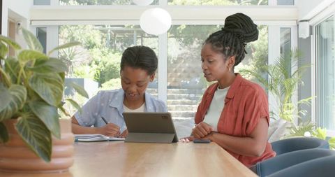 African american mom helping daughter with homework at sunlit table using tablet