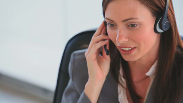 A businesswoman is using a headset, engaging in a phone call while sitting in the office. This video is ideal for representing customer support, professional communication, business consulting, and office work.