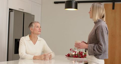 Mature and middle-aged women sharing coffee and chatting at kitchen island with holiday decor
