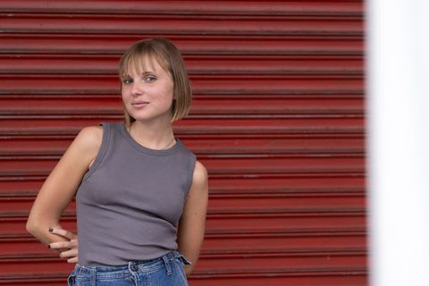 Young woman leaning against red shutter wearing gray tank top and high-waist denim