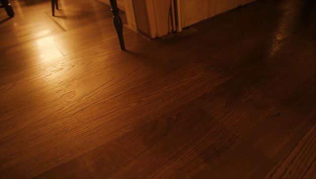 Warm dark hardwood floor reflecting amber lamp light with chair legs, scuffs, baseboard