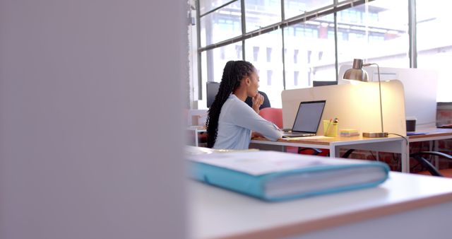 Image captures a young professional working at a laptop in a modern open office space with large windows. The ambiance is bright and organized with natural light flooding the workspace. Useful for depicting themes of productivity, modern work environments, remote work, corporate lifestyle, and concentration.