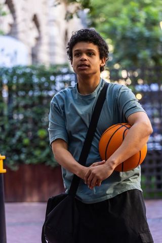 Young man holding basketball carrying duffel bag wearing teal tee urban plaza