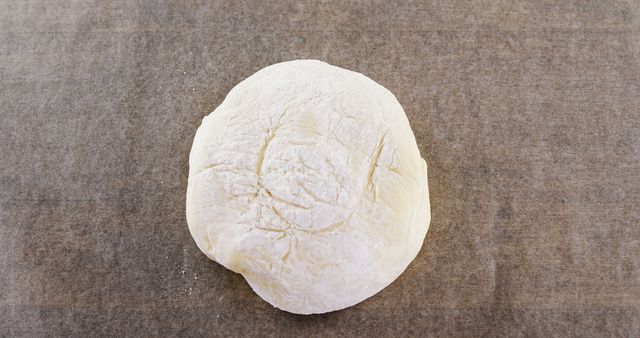 Round ball of fresh dough resting on parchment paper, perfect for illustrating baking activities, recipes, or cooking ingredients. This is ideal for use in culinary magazines, food blogs, cooking tutorials, and related content.