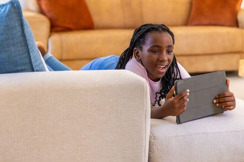 African girl lying on sofa using tablet, reacting with joy in warm beige living room