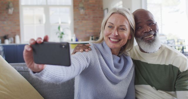 Senior couple sitting on couch in living room smiling and taking selfie with smartphone. Perfect for use in advertisements, lifestyle blogs, retirement community brochures, senior living marketing materials, and articles promoting healthy and joyful aging.
