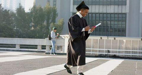 Graduate walking across campus plaza examining rolled diploma while checking phone