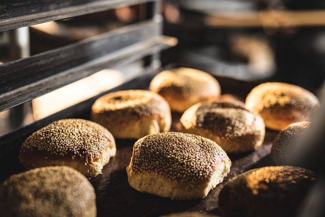 Freshly baked sesame seed breads cooling on a rack in a bakery kitchen. Ideal for use in food industry promotions, bakery advertisements, culinary blogs, and artisanal bread features. Highlights the quality and craftsmanship of homemade baking.