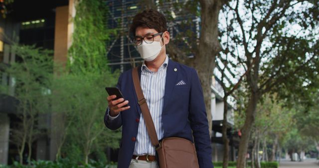 Asian businessman wearing glasses and formal attire, carrying a bag, stands on a tree-lined street. He is checking his smartphone while wearing a protective face mask, indicating attentiveness to health measures. This imagery is useful for topics related to urban lifestyle, business communication, technology use, health safety measures, and professional life.