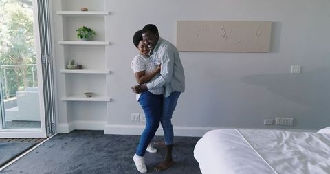 African American couple embracing and laughing in modern bedroom with balcony view