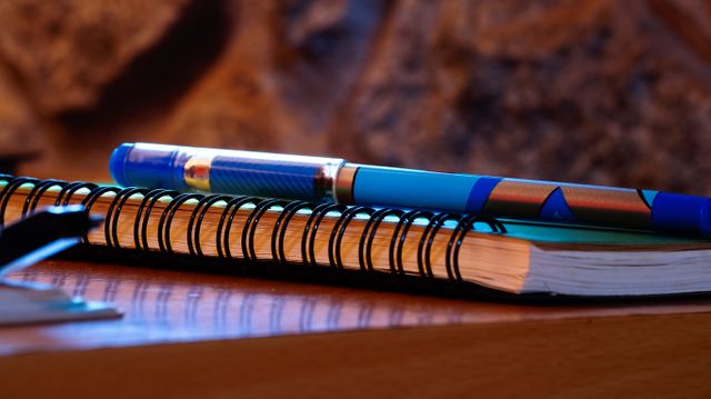 Closeup shows a pen resting on top of a spiral notebook placed on a wooden desk. Suitable for illustrating concepts related to studying, writing, taking notes, office work, education, and daily organization. Ideal for educational materials, office supply ads, productivity blogs, or workstation decorations.