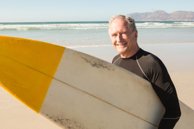 Portrait of smiling senior man with surfboard standing at beach against sky