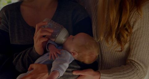 Two women feeding newborn with bottle on couch sharing gentle caregiving and bonding