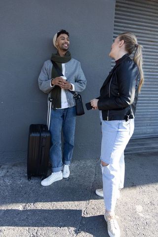 Diverse young travelers chatting on city sidewalk holding suitcase and coffee