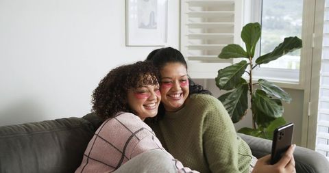 Friends taking selfie on gray couch while wearing under-eye patches and smiling together