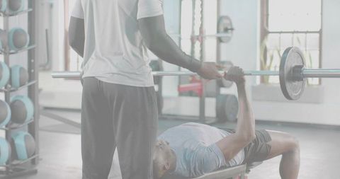 Trainer spotting bench pressing athlete lifting barbell in bright gym studio