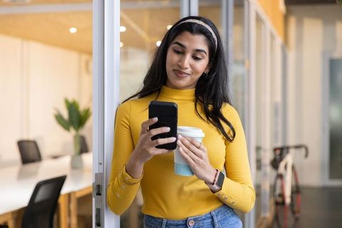 Young woman in mustard sweater checking phone holding coffee in modern coworking space