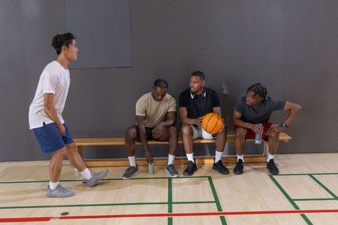 Multiracial men resting on gym bench with basketball and water bottles during team break
