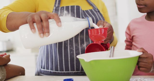 Parent and child are baking together in a kitchen, pouring milk into a red measuring cup. Child is attentive while pouring milk, learning cooking skills. They use a green mixing bowl and both wear casual clothing with aprons. Perfect for content related to family activities, bonding, teaching cooking skills, and parent-child interactions.