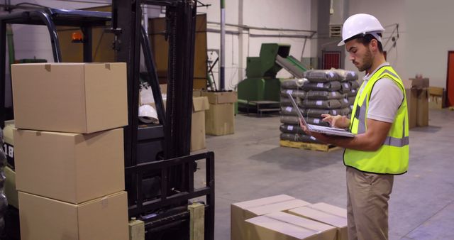 Warehouse worker in safety helmet and yellow vest using laptop for inventory management. Stacked cardboard boxes in the background along with a forklift. Useful for themes around logistics, safety protocols, warehouse management, and industrial work settings.