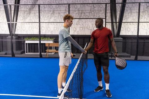 Mixed-race male players shaking hands over net on blue indoor padel court after match