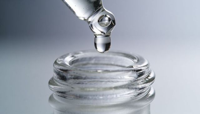 Glass dropper dropping clear serum droplet above vial neck macro laboratory skincare