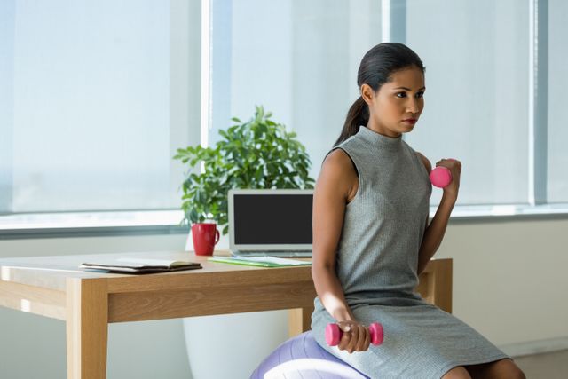 Businesswoman lifting dumbbells while sitting on a fitness ball in a modern office. Ideal for illustrating concepts of work-life balance, corporate wellness programs, and promoting a healthy lifestyle in the workplace. Suitable for use in articles, blogs, and advertisements related to office fitness, employee health, and productivity.